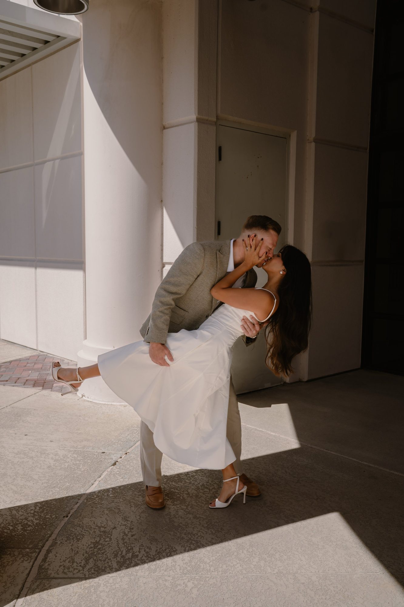 Couple kissing against a sunlit building column during wedding portraits